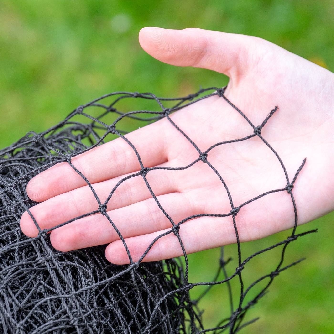 Une personne tient un rouleau ficelé de filet de protection VOSS.farming pour volailles, filet pour étang, filet pour volière, filet de protection pour rapaces (10x5m, noir) à l'extérieur, à proximité d'herbe verte et de buissons.